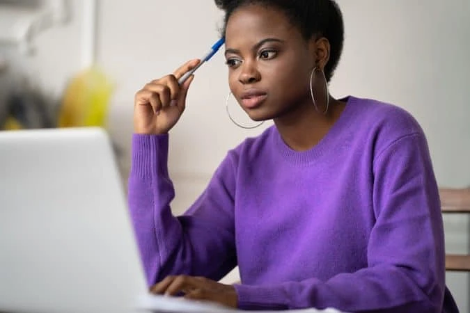 A student sits in front of their laptop wearing a purple sweater and holding a pen against their forehead