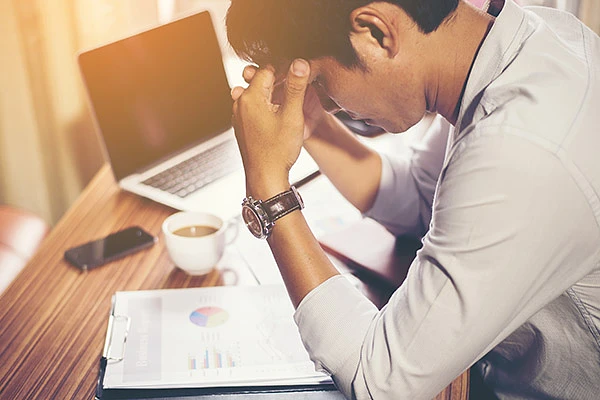 A student holds their hands on their head as the look down, feeling stressed, at a table with papers, coffee, and a laptop